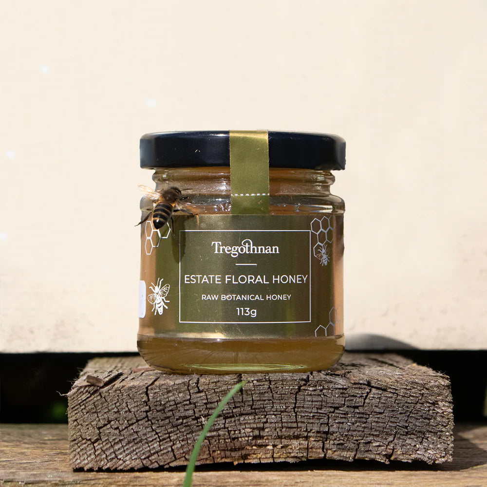 A glass jar filled with golden honey is placed on a light colored wooden surface. A bee with yellow and black stripes is perched on the rim of the jar. Text on the jar label reads "Tregothnan Estate Floral Honey" and "Raw Botanical Honey 113g