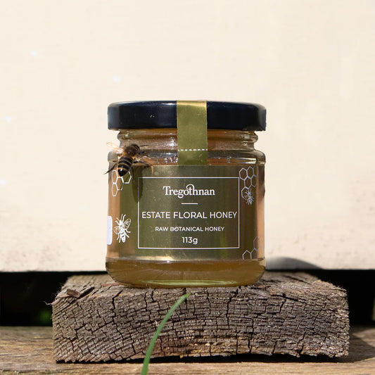 A glass jar filled with golden honey is placed on a light colored wooden surface. A bee with yellow and black stripes is perched on the rim of the jar. Text on the jar label reads "Tregothnan Estate Floral Honey" and "Raw Botanical Honey 113g