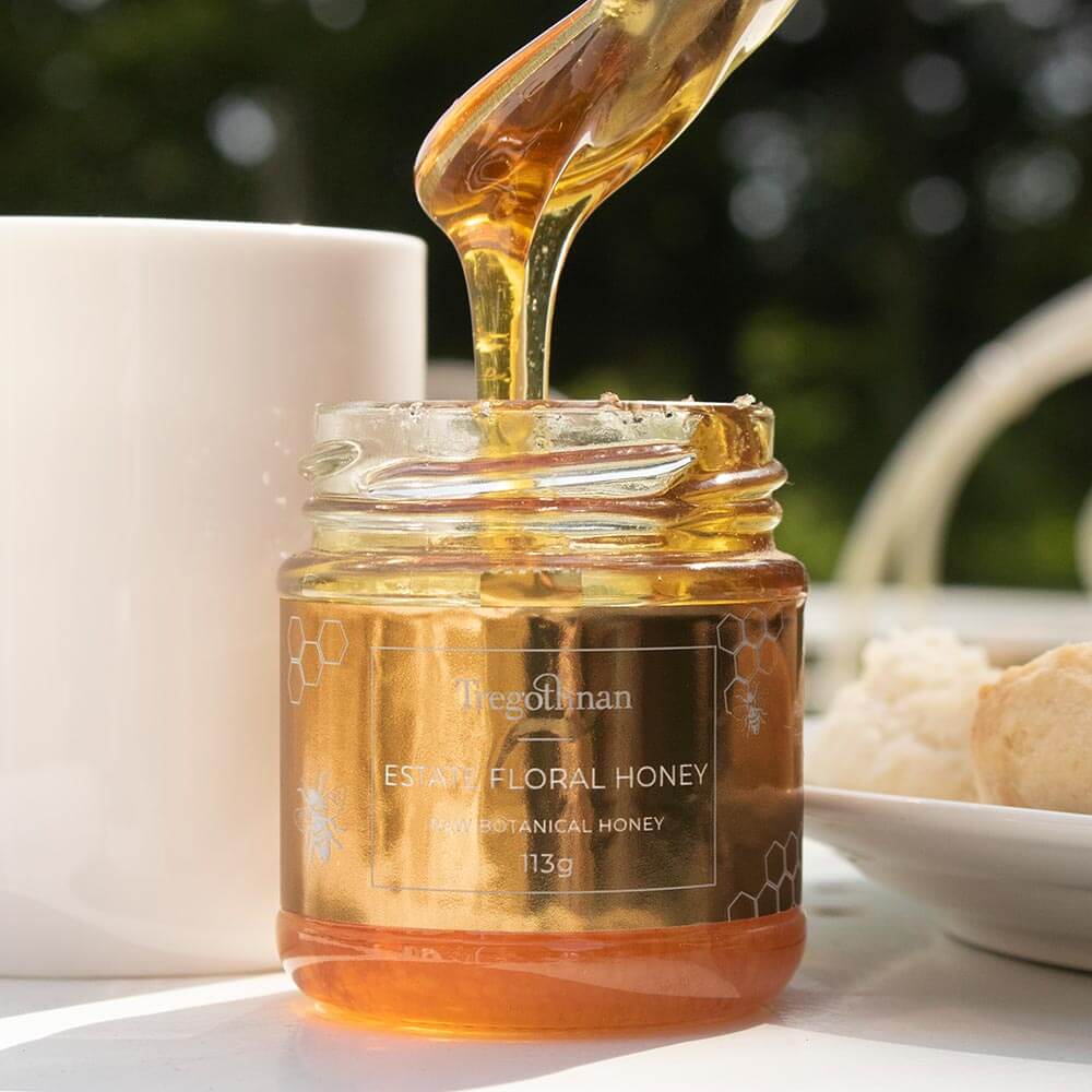 Close up of a jar of the Cornish Tregothnan Estate Floral honey with a spoon in it. The jar has a label that says "Tregothnan Estate Floral Honey, Raw Botanical Honey, 113g".