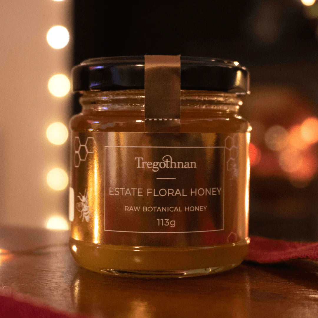 A jar of the Cornish Tregothnan Estate Floral Honey, a type of raw botanical honey, sits on a table. The honey is a golden colour and appears viscous. The 113g glass jar has a gold label with white text. 